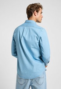 Ljusblå denimskjorta med långa ärmar, knappstängning och veckdetalj bak. Tyget har en mjuk, strukturerad finish.