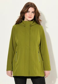 Giacca verde con cappuccio e tasche laterali con zip, caratterizzata da una texture morbida e un design dal fit rilassato. Il tessuto appare leggero e liscio.