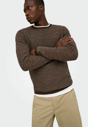Homme les bras croisés portant un pull marron texturé sur une chemise blanche et un pantalon beige, regardant vers sa gauche sur un fond uni.