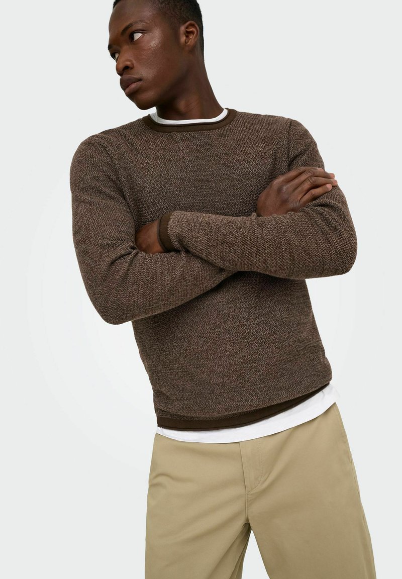 Homme les bras croisés portant un pull marron texturé sur une chemise blanche et un pantalon beige, regardant vers sa gauche sur un fond uni.