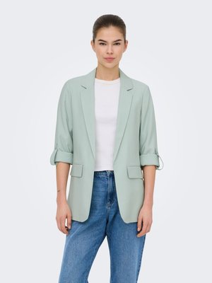 Jonge vrouw draagt een lichtgroene blazer met opgerolde mouwen, wit T-shirt en blauwe spijkerbroek, staand tegen een effen witte achtergrond.