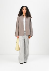 Light brown button-up shirt with wide sleeves, worn over a white top. Paired with grey striped wide-leg pants and holding a yellow bag.