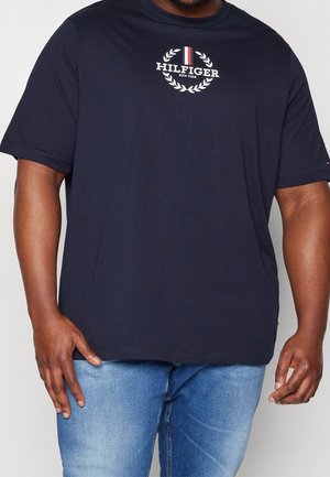 Hombre vistiendo camiseta azul marino de Tommy Hilfiger con logo blanco y vaqueros azul claro sobre fondo liso.