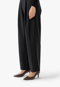 Ženska oblečena v črne široke hlače in rjave čevlje s špicastim prstom, stoji z eno roko vidno, ki prikazuje poročne prstane.