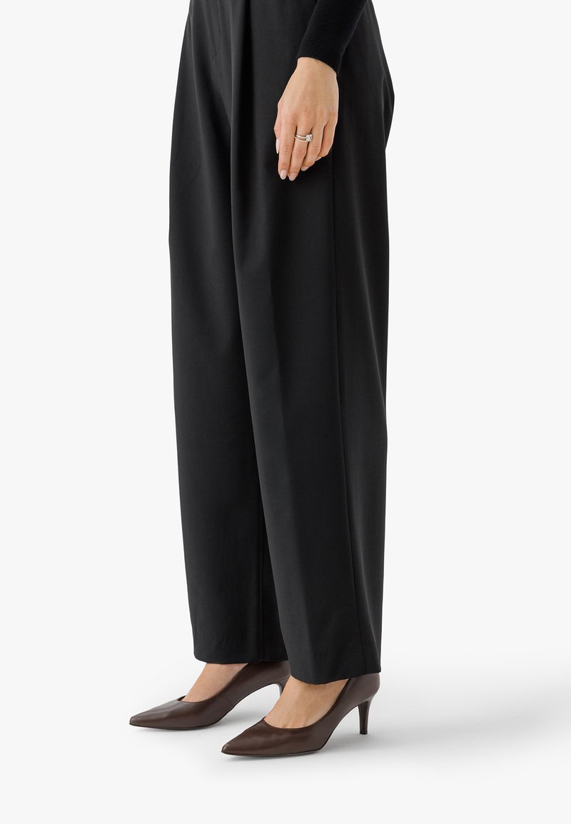 Ženska oblečena v črne široke hlače in rjave čevlje s špicastim prstom, stoji z eno roko vidno, ki prikazuje poročne prstane.