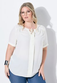 Weißes Blusenshirt mit kurzen Ärmeln, einem gebundenen Halsausschnitt und einem fließenden Design. Kombiniert mit blauen Jeans und einem silbernen Armreif.