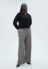 Schwarzer Pullover mit Kragen, graue weit geschnittene Hose und Schuhe aus Lackleder. Das Model steht mit den Händen in der Hüfte vor einem hellen Hintergrund.