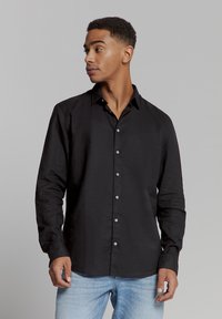 Zwarte langemouwen shirt van textuurstof, met een button-down kraag, knopen aan de voorkant en een relaxte pasvorm. Gepaard met lichtblauwe jeans.