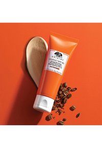 Oransje tube med Origins Ginzing SPF 40 fuktighetskrem med hvit lokk, sammen med kaffebønner på en lys orange bakgrunn.