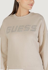 Felpa beige con scollo e polsini a coste. Presenta un logo "GUESS" in strass argentati di grande rilievo sul davanti.