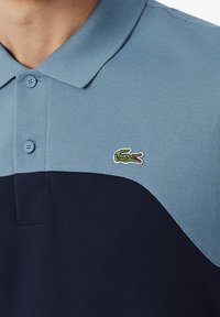 Short-sleeve polo shirt featuring light blue and dark navy color blocks, textured fabric, and a small embroidered crocodile logo.