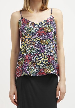 Femme portant un haut sans manches à imprimé floral avec de fines bretelles et une jupe noire, debout devant un fond blanc uni.