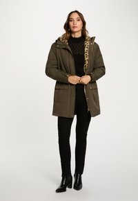 Donna che indossa un parka verde con foderatura in pelliccia sintetica leopardata, un maglione nero a fantasia, pantaloni neri e stivali neri con tacco, in piedi davanti a uno sfondo bianco.