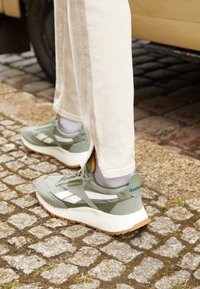 Green athletic shoes with white accents, suede and mesh materials, featuring a thick white sole and a branded heel tab. Worn with light-colored pants.