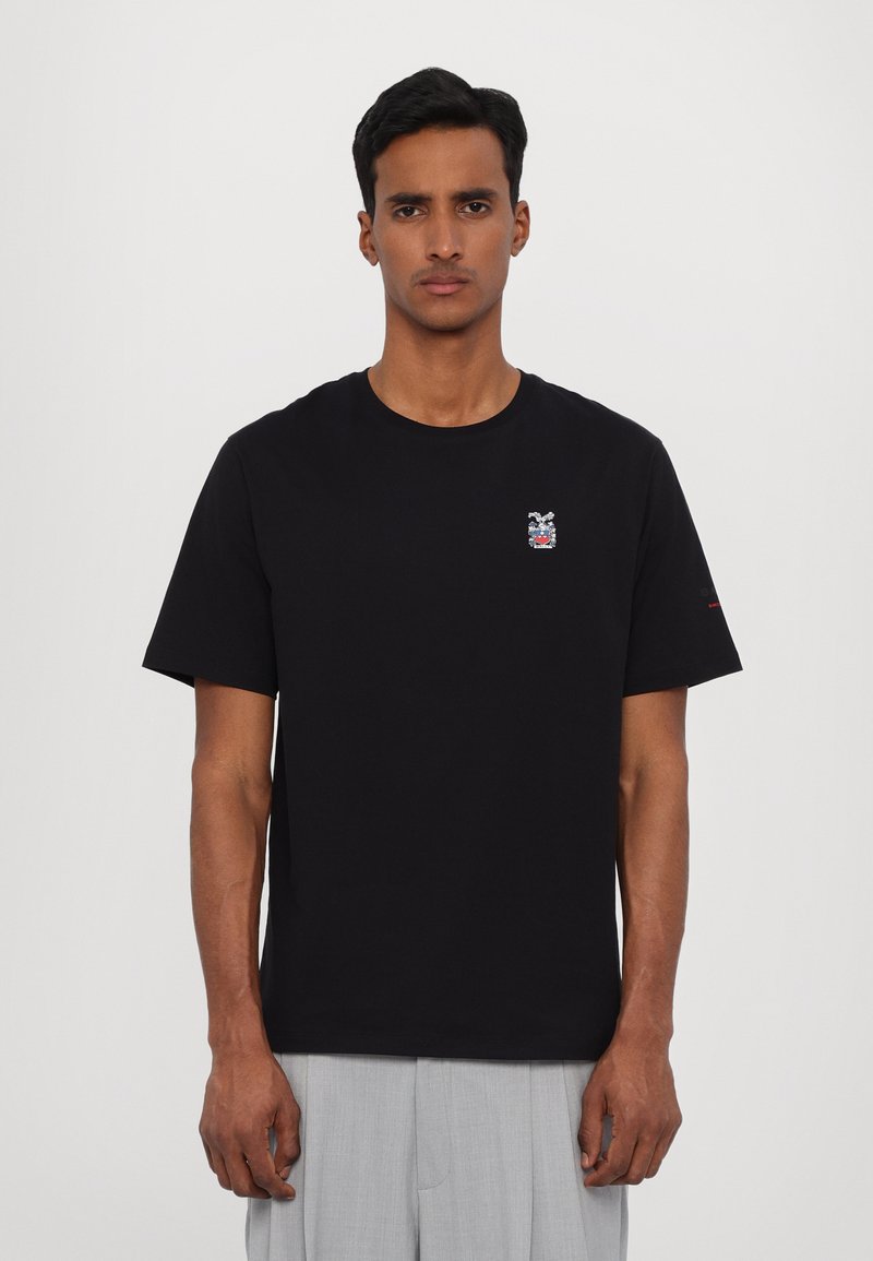 T-shirt en coton noir avec un col rond, des manches courtes, comportant un petit motif brodé sur la poitrine et un logo discret sur la manche.