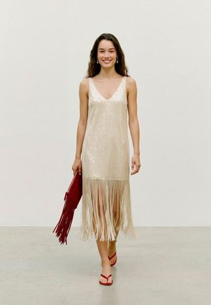 Une femme souriante défile sur un podium dans une robe crème à sequins et à longues franges, avec des sandales rouges et une pochette rouge à franges.