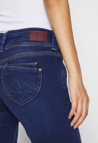 Tmavě modré džíny z denimu mají zadní kapsu s šikmým designem a hnědou koženou záplatou v oblasti pasu. Švy jsou viditelné podél okrajů.