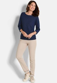 Marineblauer Strickpullover mit strukturiertem Muster, Rundhalsausschnitt und Dreiviertelärmeln; kombiniert mit beigen, figurbetonten Hosen und hellen Schuhen.