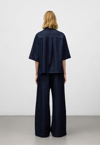 Camicia in denim scuro a maniche corte e vestibilità ampia, abbinata a pantaloni in denim scuro a gamba larga. La vista sul retro mostra dettagli di cucitura discreti.
