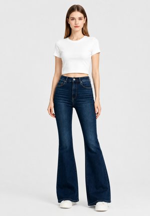 Young woman standing wearing a white cropped t-shirt, high-waisted dark blue flared jeans, and white sneakers on a plain light background.