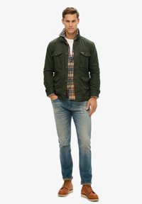 Chaqueta de algodón color verde oliva con dos bolsillos frontales, superpuesta sobre una camisa de cuadros. Unos vaqueros azul claro y unas botas de cuero marrón completan el atuendo.