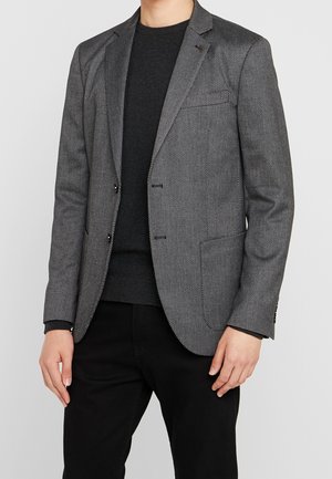Homme portant un blazer gris texturé ajusté sur un pull noir et un pantalon noir, debout les mains détendues le long du corps.