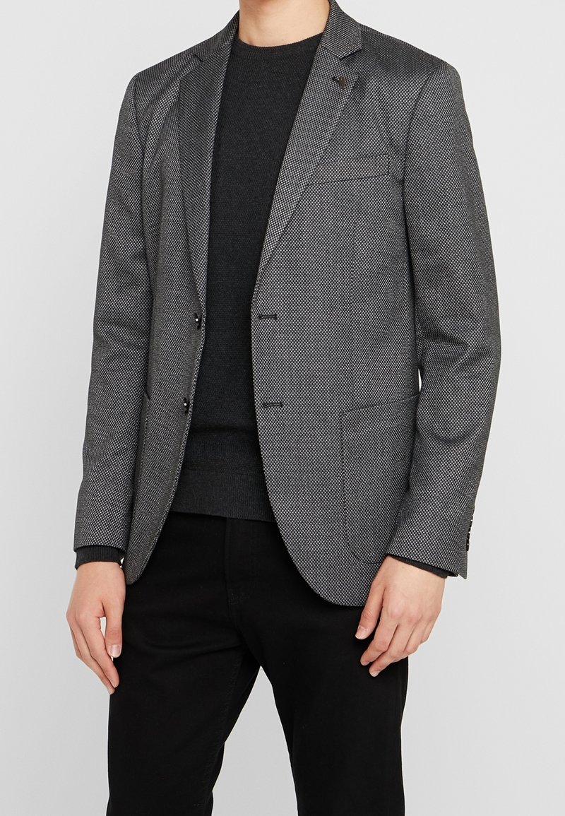 Homme portant un blazer gris texturé ajusté sur un pull noir et un pantalon noir, debout les mains détendues le long du corps.