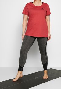 Camiseta roja de manga corta con un logo negro, combinada con leggings gris oscuro que presentan acentos negros y un acabado texturizado.