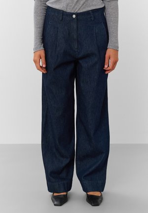 Personne portant un jean bleu foncé à jambes larges, un haut gris à manches longues et des chaussures noires pointues, debout devant un fond uni.