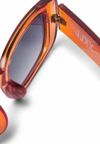 Oranžne prozorčaste sončne očala z debelimi okvirji in temnimi lečami, ki imajo zlat tečaj in belo logo na nogici.