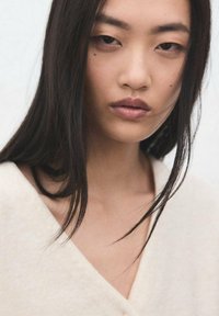 Retrato en primer plano de una persona con cabello largo, lacio y negro, maquillaje neutro y una prenda suave de color crema con diseño de escote en V.