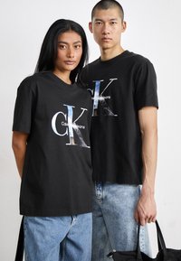 Crne pamučne majice kratkih rukava sadrže veliki reflektirajući "CK" logo i tekst "Calvin Klein Jeans". Nosive su s svijetlom plavom trapericom.
