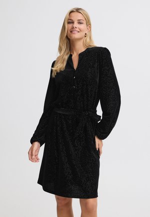 Femme aux longs cheveux blonds portant une robe noire texturée à manches longues avec des boutons et une ceinture nouée à la taille, debout et souriante.