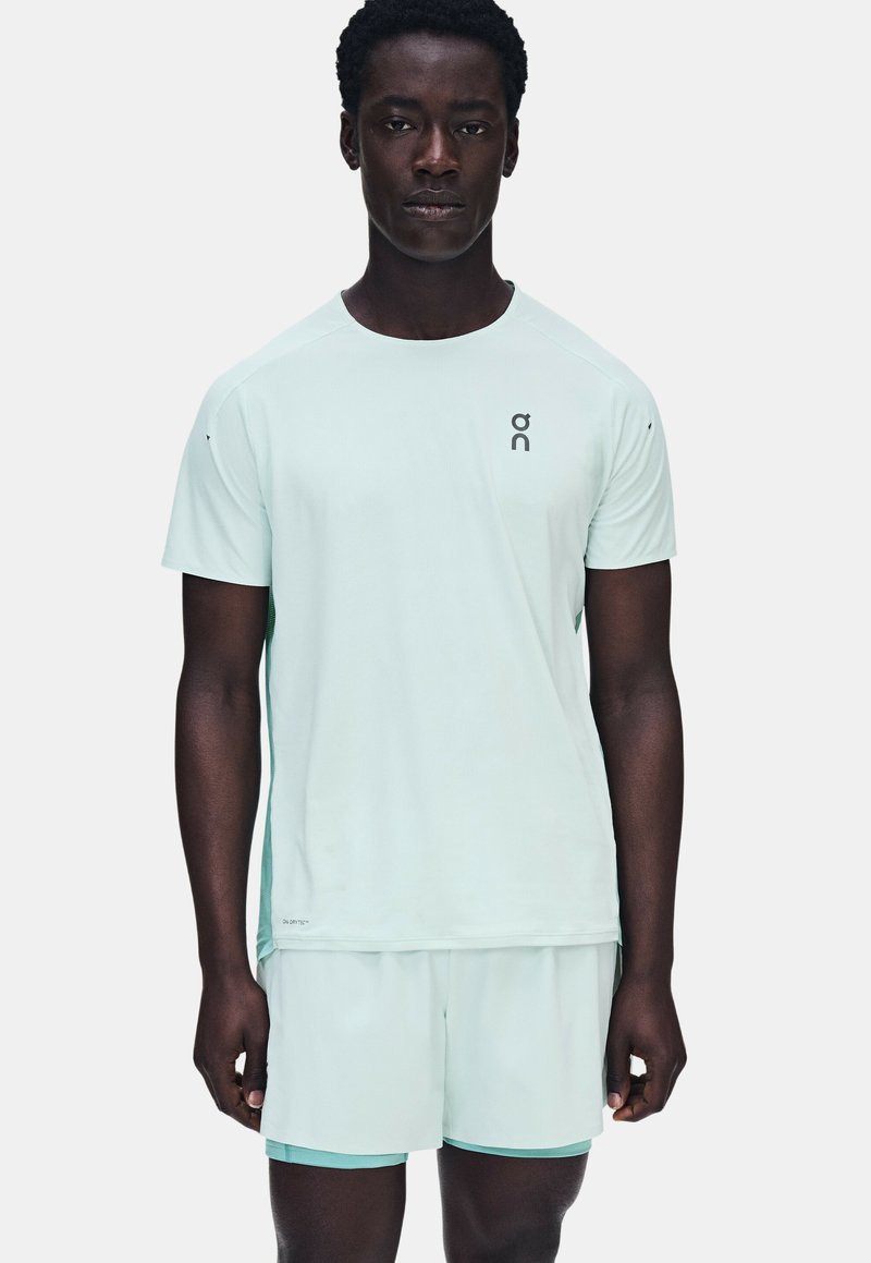 Homme portant un t-shirt de sport bleu clair à manches courtes et un short assorti, debout devant un fond uni.