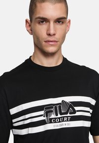 Schwarzes T-Shirt mit Rundhalsausschnitt, versehen mit weißen horizontalen Streifen und einem auffälligen FILA-Logo, das mittig auf der Vorderseite platziert ist. Aus Baumwollmaterial.