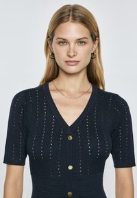 Cardigan a maglia blu navy con maniche corte, dotato di scollo a V, coste orizzontali e chiusura con bottoni. Progettato con dettagli a maglia aperta in tutto il capo.