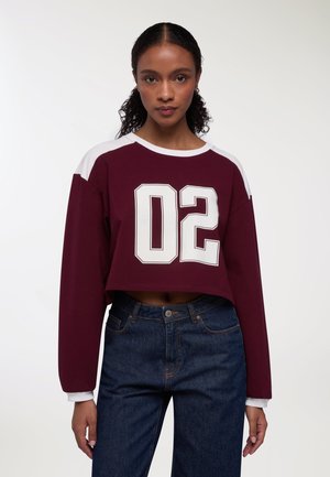 Felpa corta bordeaux con maniche bianche e grande grafica "02". Materiale morbido, vestibilità comoda e polsini a costine. Indossata con jeans di denim scuro.