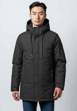 Homme portant une veste d'hiver noire à capuche avec des fermetures éclair et des boutons, se tenant contre un fond clair.