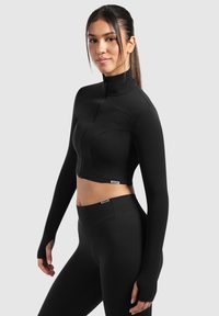 Schwarze, kurz geschnittene Trainingsjacke mit langen Ärmeln, hohem Kragen und Reißverschluss vorne. Glatte Textur mit subtilen Nahtdetails. Kombiniert mit passenden Leggings.