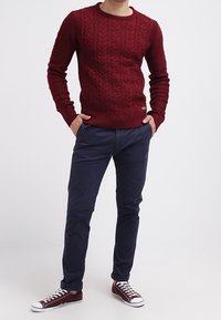 Mann trägt einen roten Zopfstrickpullover, marineblaue Hose und dunkelrote Turnschuhe, steht mit den Händen in den Taschen vor einem einfarbigen Hintergrund.