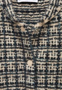 Veste en laine texturée avec un motif à carreaux noir, beige et bleu, équipée de boutons-pression et d’un col.