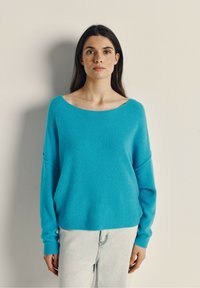 Turkis oversized sweater med bred halsudskæring og sænkede skuldre, lavet af et blødt, struktureret materiale. Båret med lyse denimjeans.