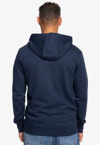 Námořnická modrá mikina s kapucí na šňůrku, žebrovanými manžetami a spodním lemem; vyrobena z měkké tkaniny; jednoduchý design bez viditelných vzorů nebo loga.