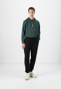 Mørkegrønn hettegenser med logo, frontlomme og snor. Svart joggebukser og kremfargede sneakers. Ensfarget design, avslappet passform.