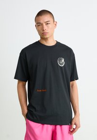 Mand med kort hår iført sort T-shirt med hvidt cirkulært design og orange "Texas Fossils" tekst, parret med lyserøde shorts.
