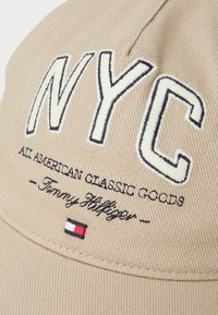 Beige baseballcaps med broderte "NYC" i hvit og mørk blå, med teksten "ALL AMERICAN CLASSIC GOODS" og et lite logo-patch.