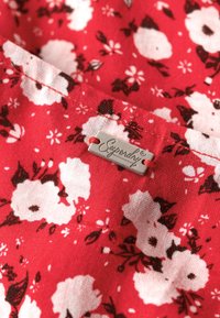Roter Stoff mit weißem Blumenmuster. Enthält ein silbernes Etikett mit dem "Superdry"-Logo, akzentuiert mit roten Nähten. Weiche Textur.