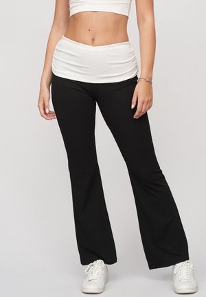 Frau trägt schwarze ausgestellte Hose, ein weißes kurzes Top, weiße Turnschuhe und ein Armband, steht vor einem einfarbigen Hintergrund.