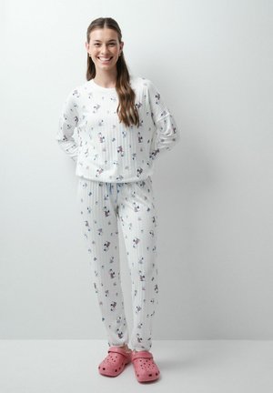 Jeune femme souriante portant un pyjama blanc avec de petits motifs de dessins animés et des chaussures roses à enfiler sur un fond blanc uni.