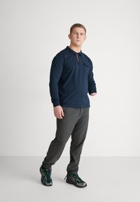 Granatowa koszulka polo z długim rękawem i kieszonką na piersi, w zestawieniu z szarymi spodniami i czarno-zielonymi trampkami. Casualowy, komfortowy krój.
